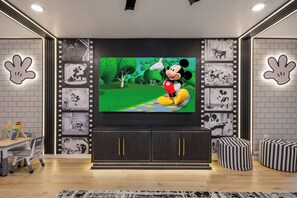 A modern living room with a large flat-screen TV, a dark wooden entertainment unit, and Mickey Mouse-themed wall decorations. - Disney Dream Home Mario, Frozen & Minions (Davenport)
