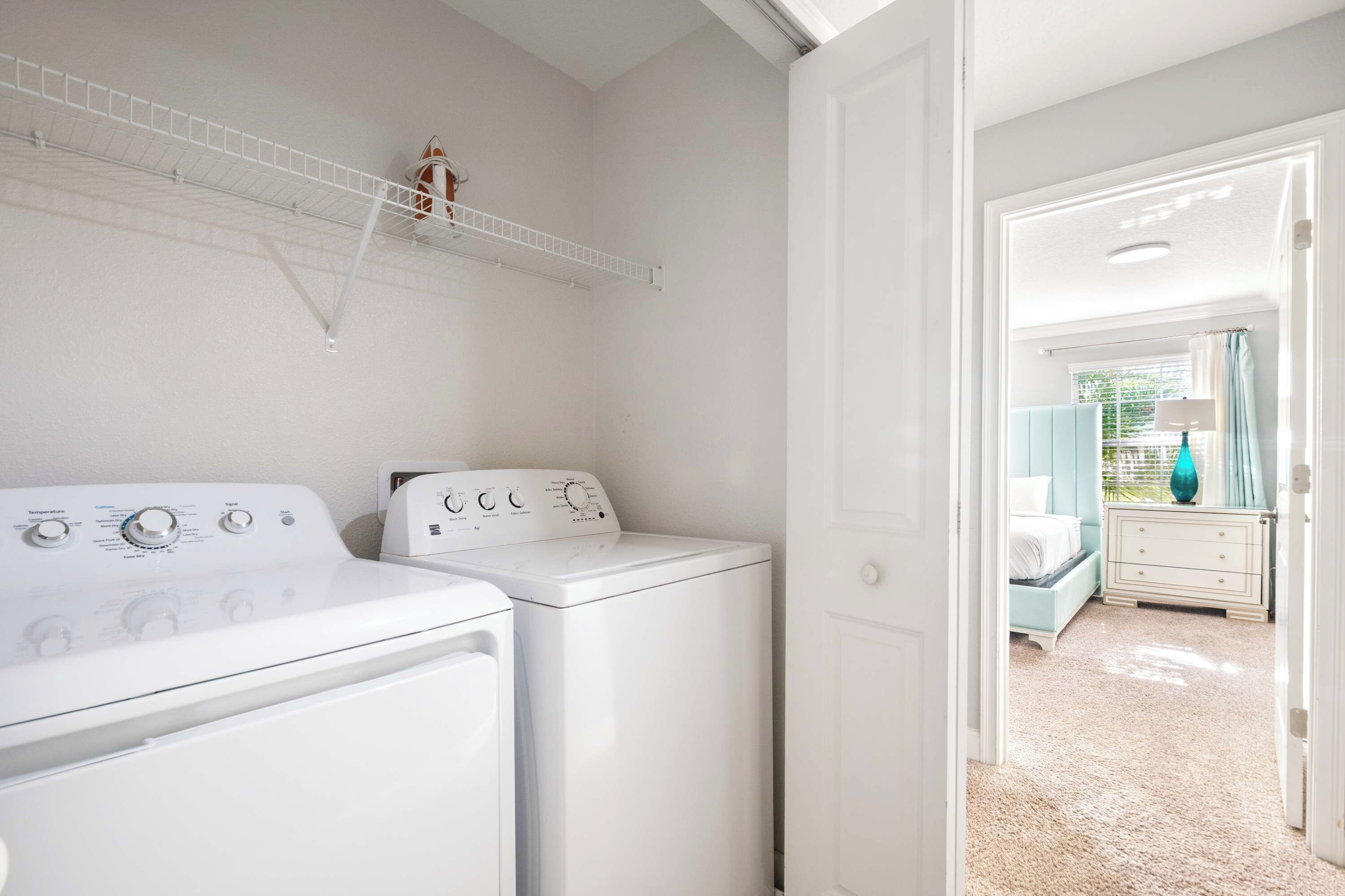 A laundry area with two white washing machines, a white shelf above, and a door leading to a bedroom with a bed and dresser.