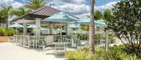 Un espace extérieur aménagé en terrasse, avec plusieurs tables et chaises, un coin salon abrité et une piscine en arrière-plan.