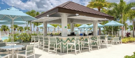 A covered outdoor seating area with tables and chairs, a pool in the background, and palm trees.