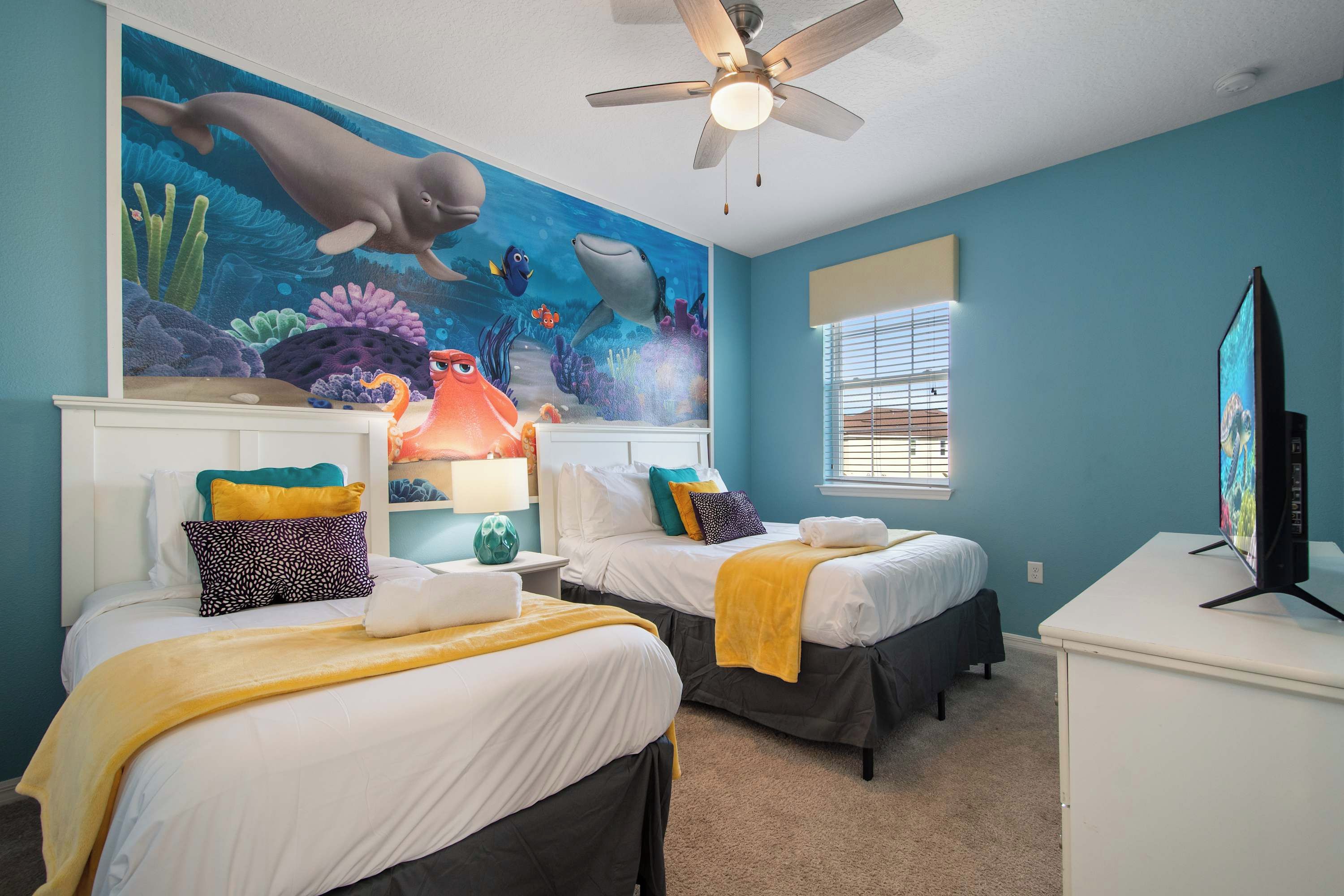 A room with two beds, a ceiling fan, a mural of marine life, and a television on a desk.