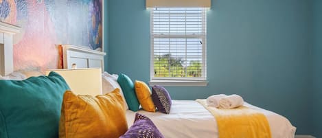 A bedroom with two beds, colorful pillows, a painting on the wall, and a window with blinds.