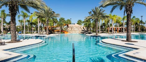 A large, clear swimming pool surrounded by palm trees and lounge chairs, with a rocky hill in the background.