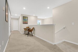 A hallway with a carpeted floor, a dining table with chairs, a framed picture on the wall, and a window with a view of the sky. - Cozy 5BR Solterra Home w/ Private Pool (Davenport)