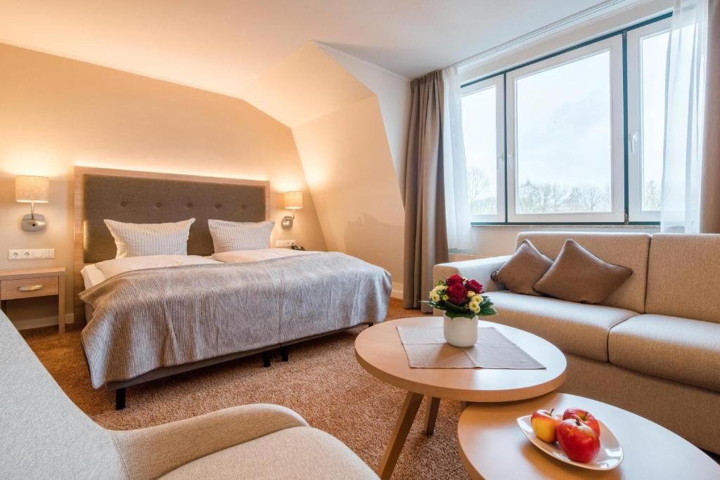 A hotel room with a bed, a sofa, a round table with a plate of fruit, and a window with curtains.
