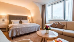 A hotel room with a bed, a sofa, a round table with a plate of fruit, and a window with curtains. - Ringhotel Altes Zollhaus (Wangerland)