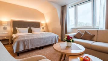 A hotel room with a bed, a sofa, a round table with a plate of fruit, and a window with curtains.