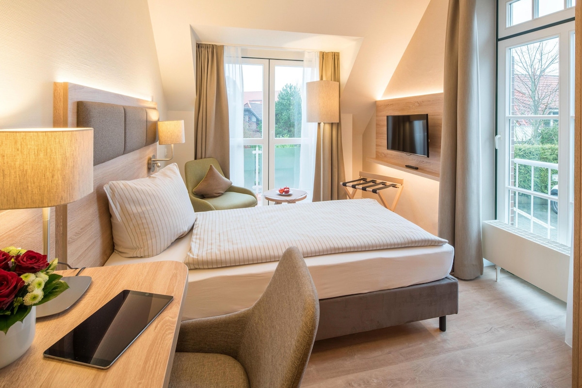 Habitación de hotel con cama, escritorio con una tablet, silla, televisor montado en la pared y ventana con cortinas.