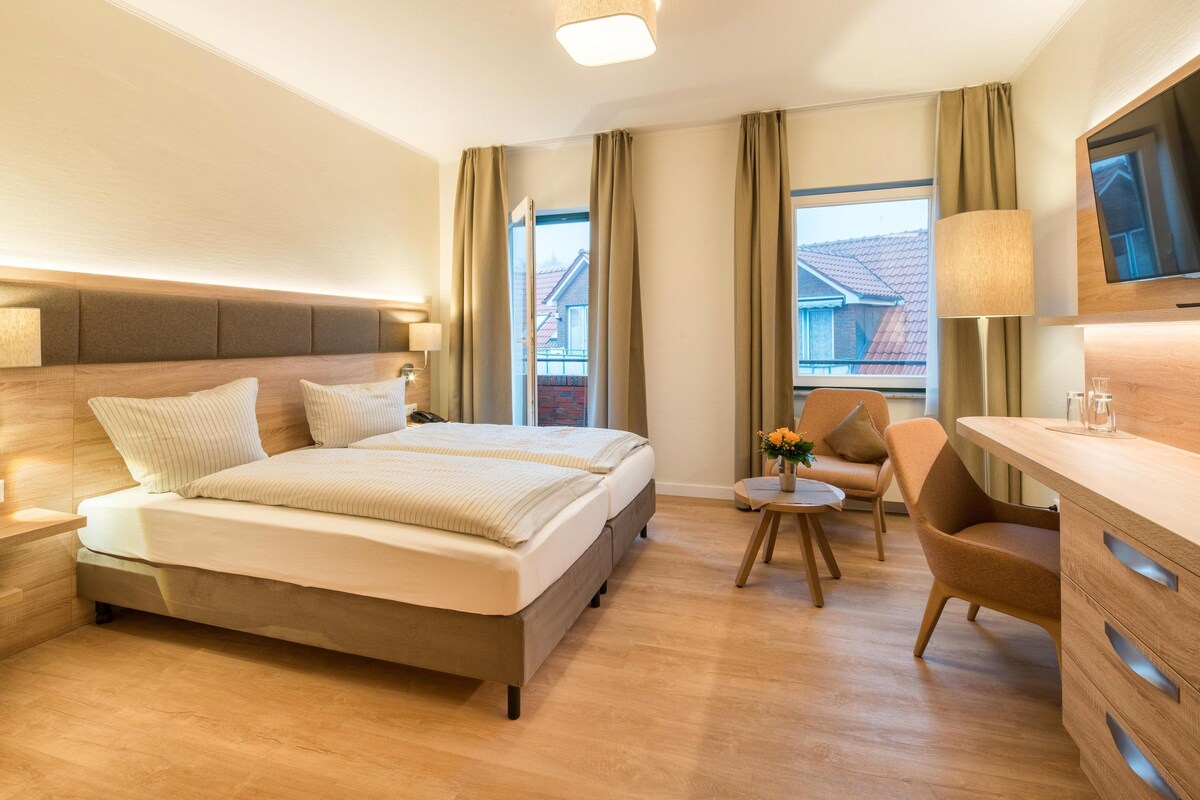 Habitación de hotel con cama, mesitas de noche, escritorio, silla, televisor y ventana con cortinas.
