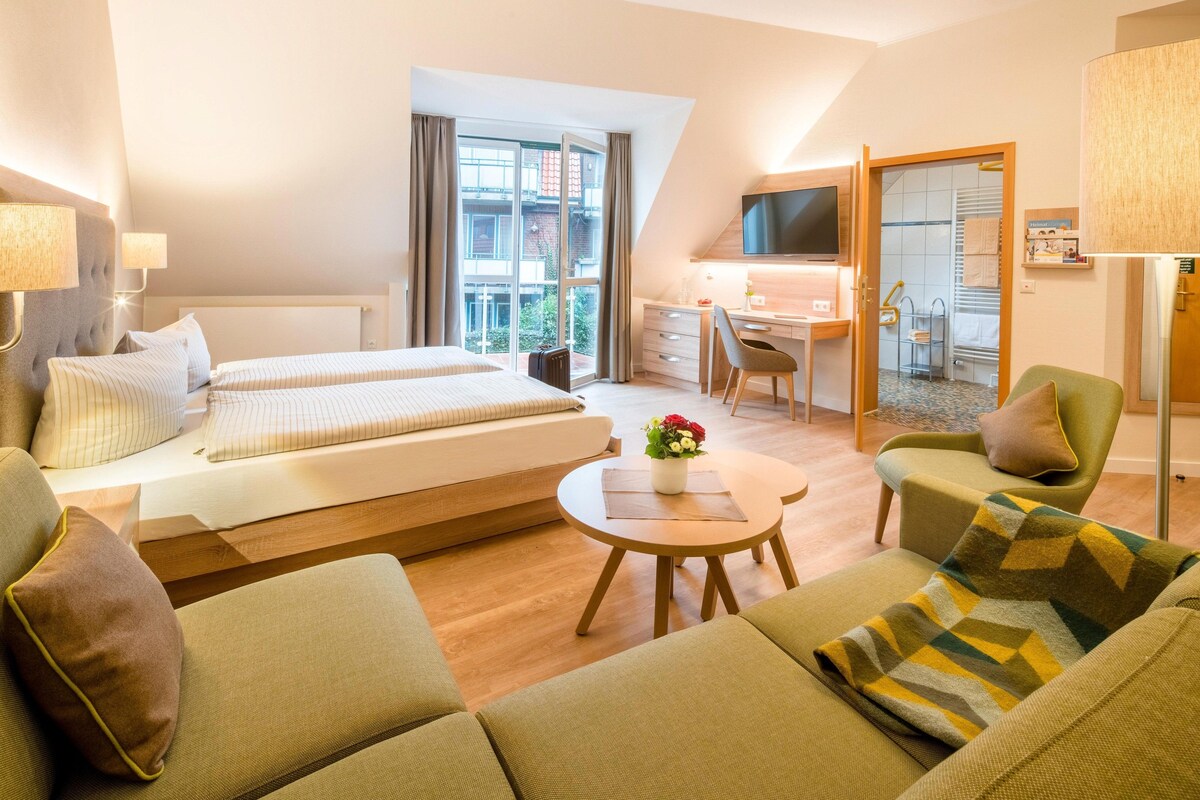 Habitación de hotel con dos camas, un escritorio, una silla, una mesita con un florero y un televisor.