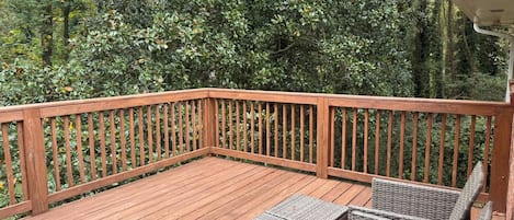 A wooden deck with a black table and wicker chairs, surrounded by dense foliage.