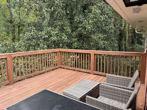 Een houten terras met een zwarte tafel en rieten stoelen, omgeven door weelderige begroeiing.
