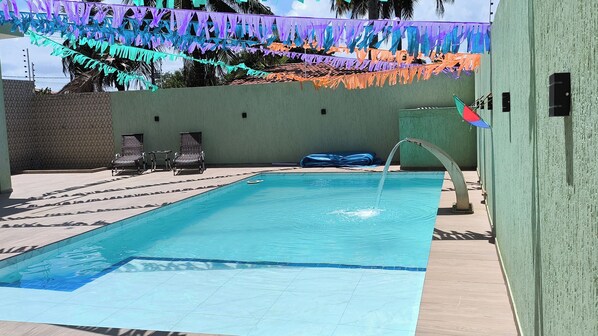 Ein Schwimmbecken mit einer Springbrunnen, umgeben von Liegestühlen und einer Wand mit bunten Flaggen.
