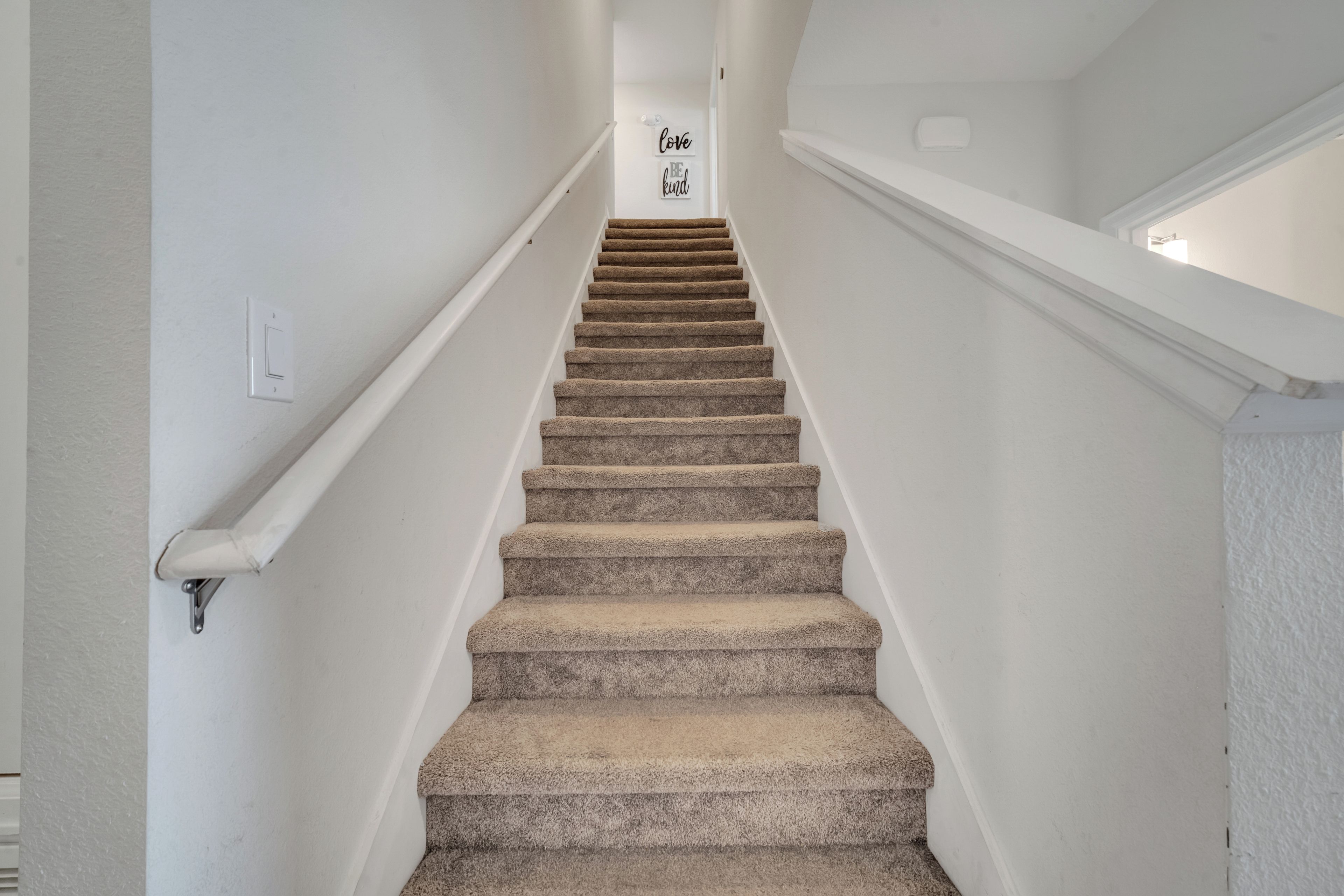 Eine Treppe mit Teppichstufen und weißen Wänden, die zu einer Zwischenebene führt, auf der ein Schild mit der Aufschrift 'love be kind' steht.