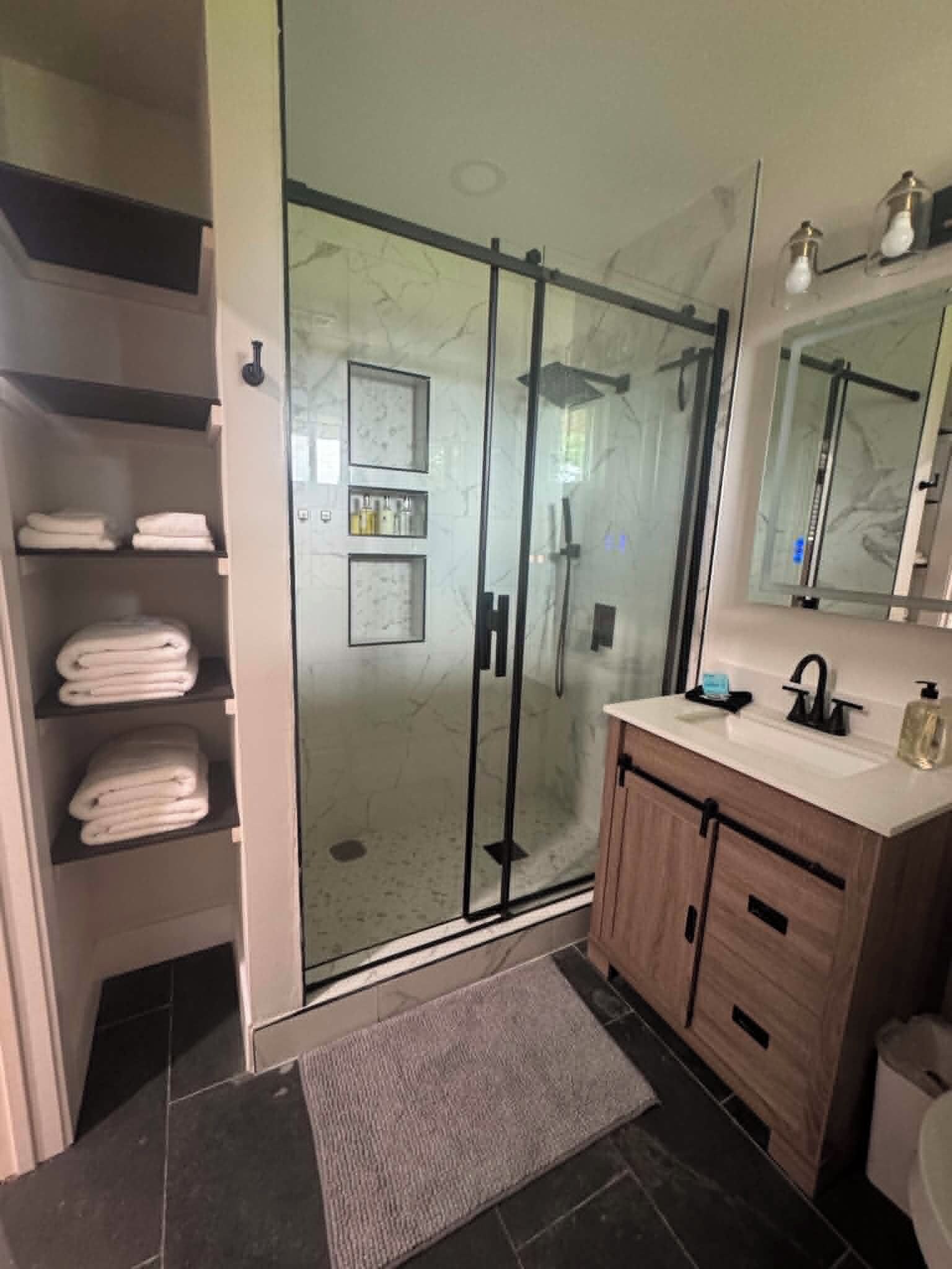 Ein modernes Badezimmer mit einer Duschkabine aus Glas, einem Holzbadezimmermöbel und einem eingebauten Regal mit gefalteten Handtüchern.
