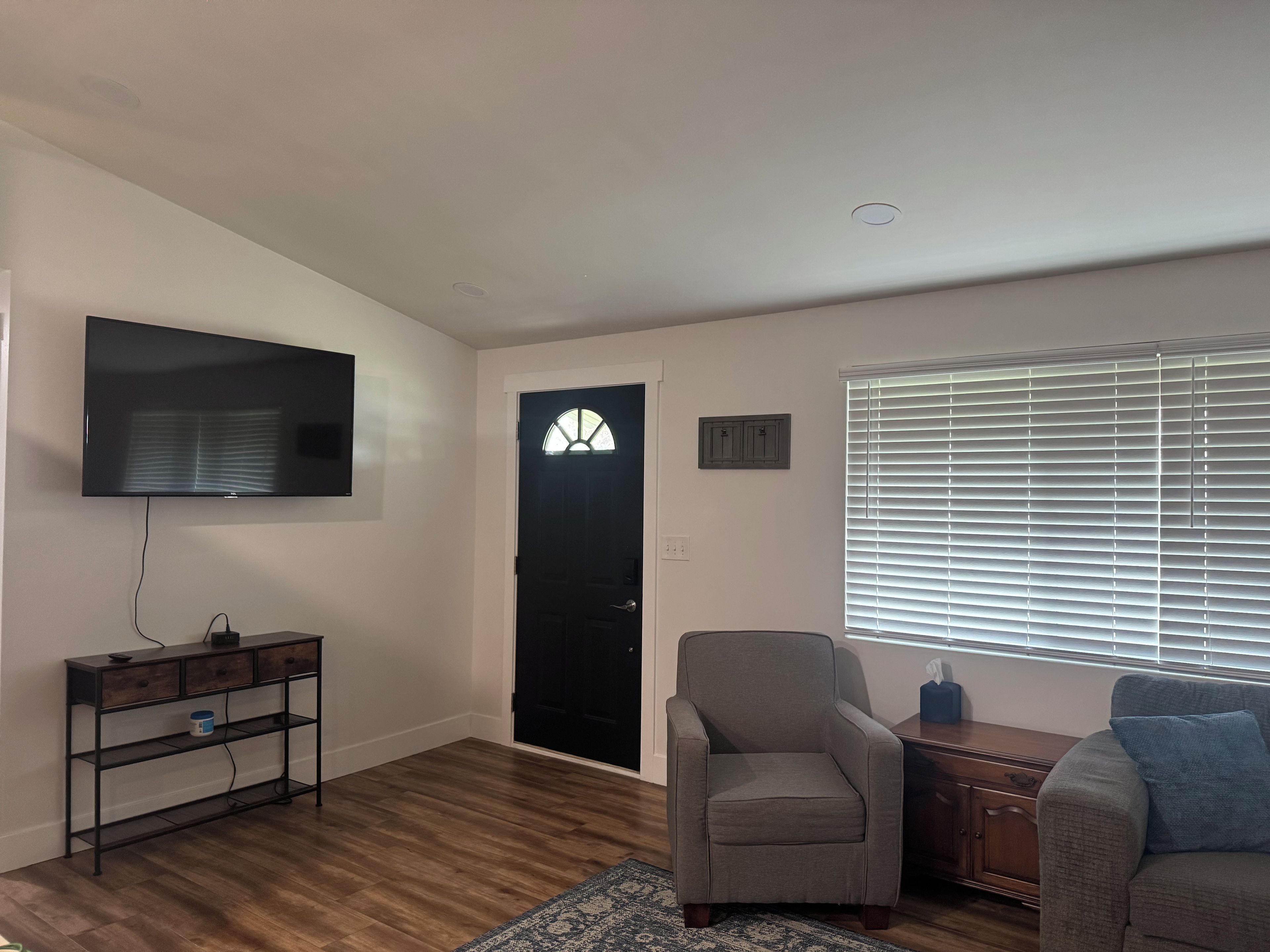 Uma sala de estar com televisão de tela plana, sofá, poltrona, piso de madeira, porta com janela e uma janela com persianas.