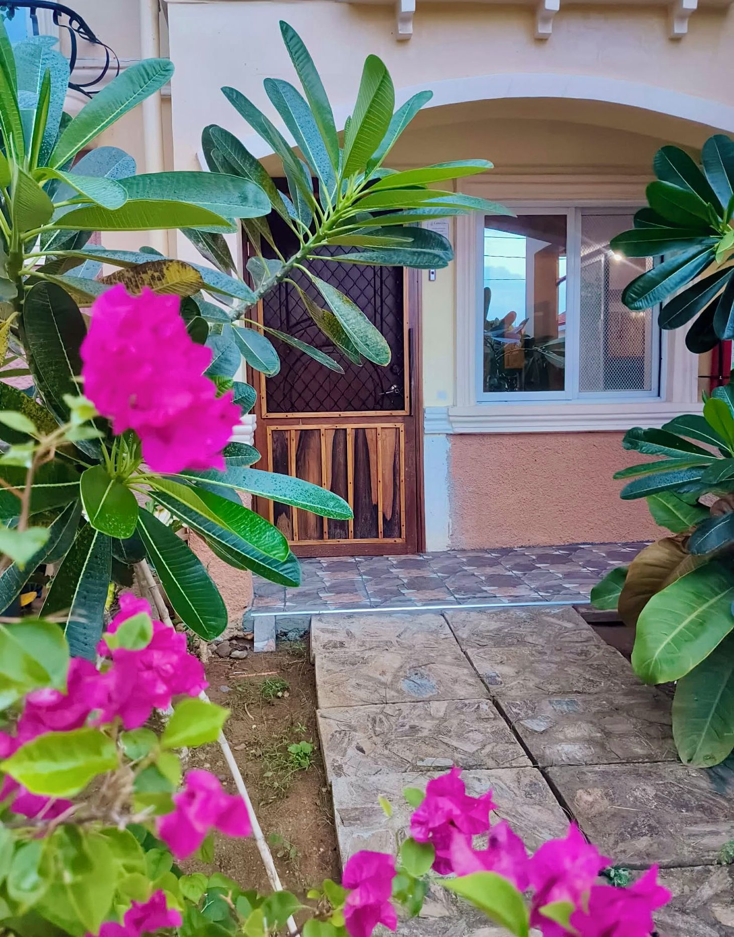 Una entrada pavimentada con una puerta de madera, rodeada de abundante follaje verde y coloridas flores rosadas.