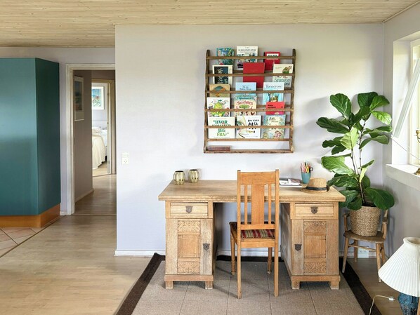 A room with a wooden desk, a chair, a bookshelf with books, a large plant, and a lamp.