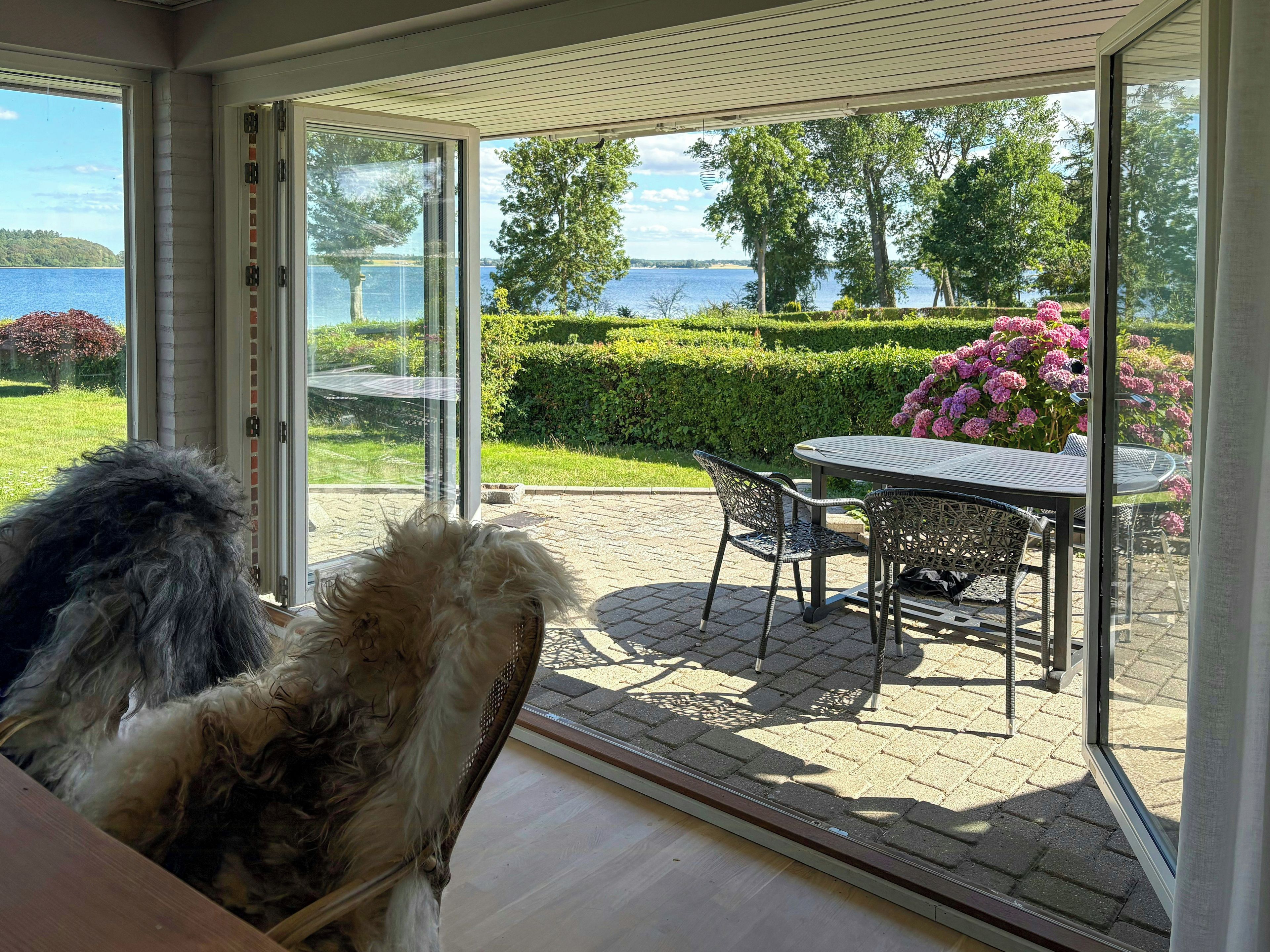 A glass-enclosed patio with a table and chairs, overlooking a landscaped garden and a body of water.