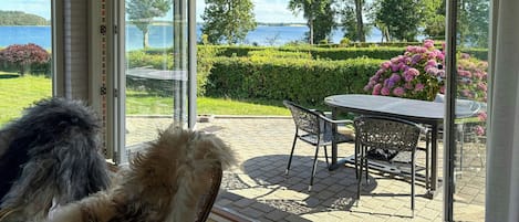 A glass-enclosed patio with a table and chairs, overlooking a landscaped garden and a body of water.