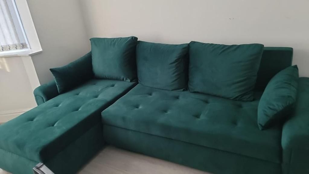 A green sectional sofa with a chaise lounge in a room with a plain wall and a window.