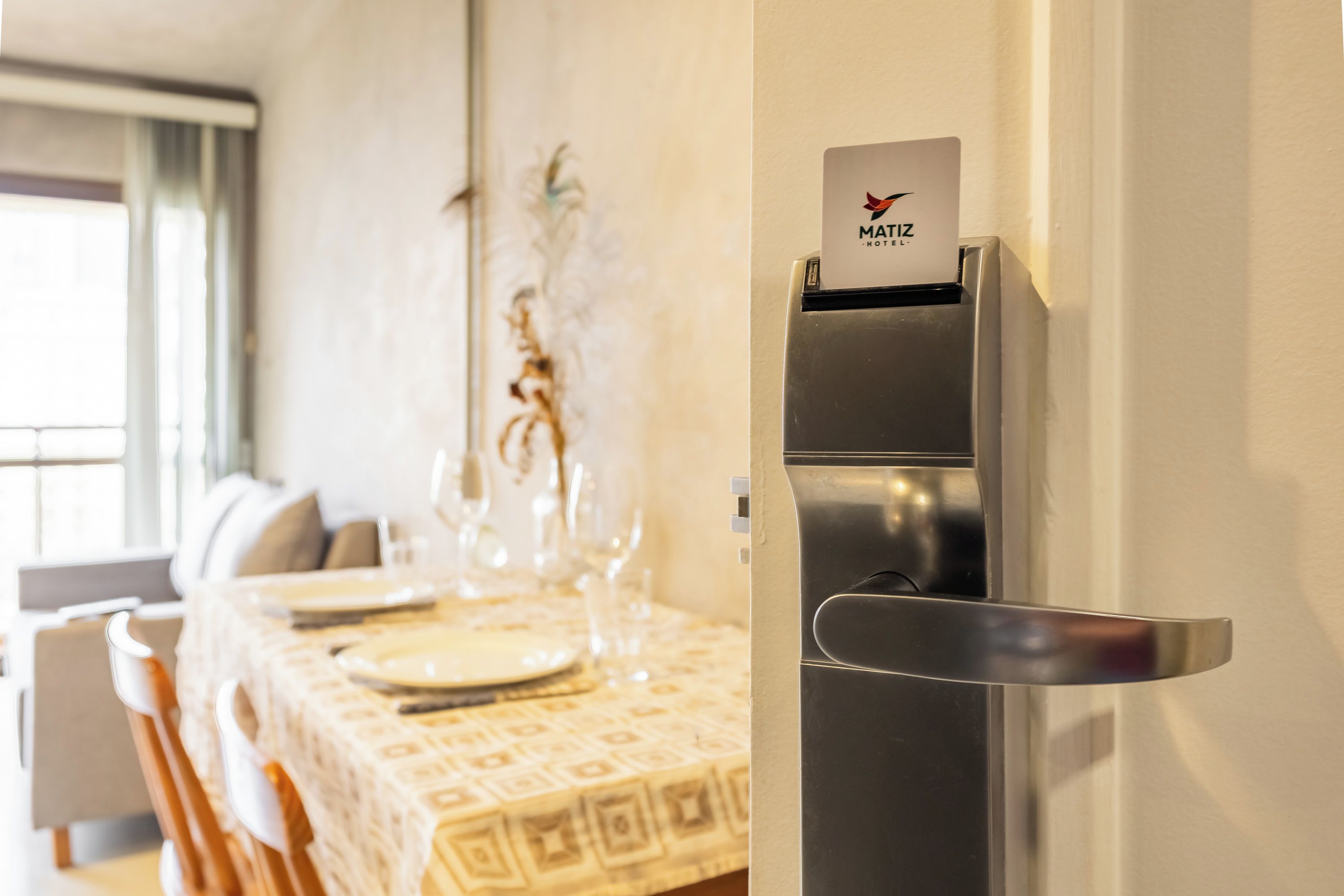 A modern hotel room with a dining area featuring a table set for four, a minibar, and a door with a hotel name sign.