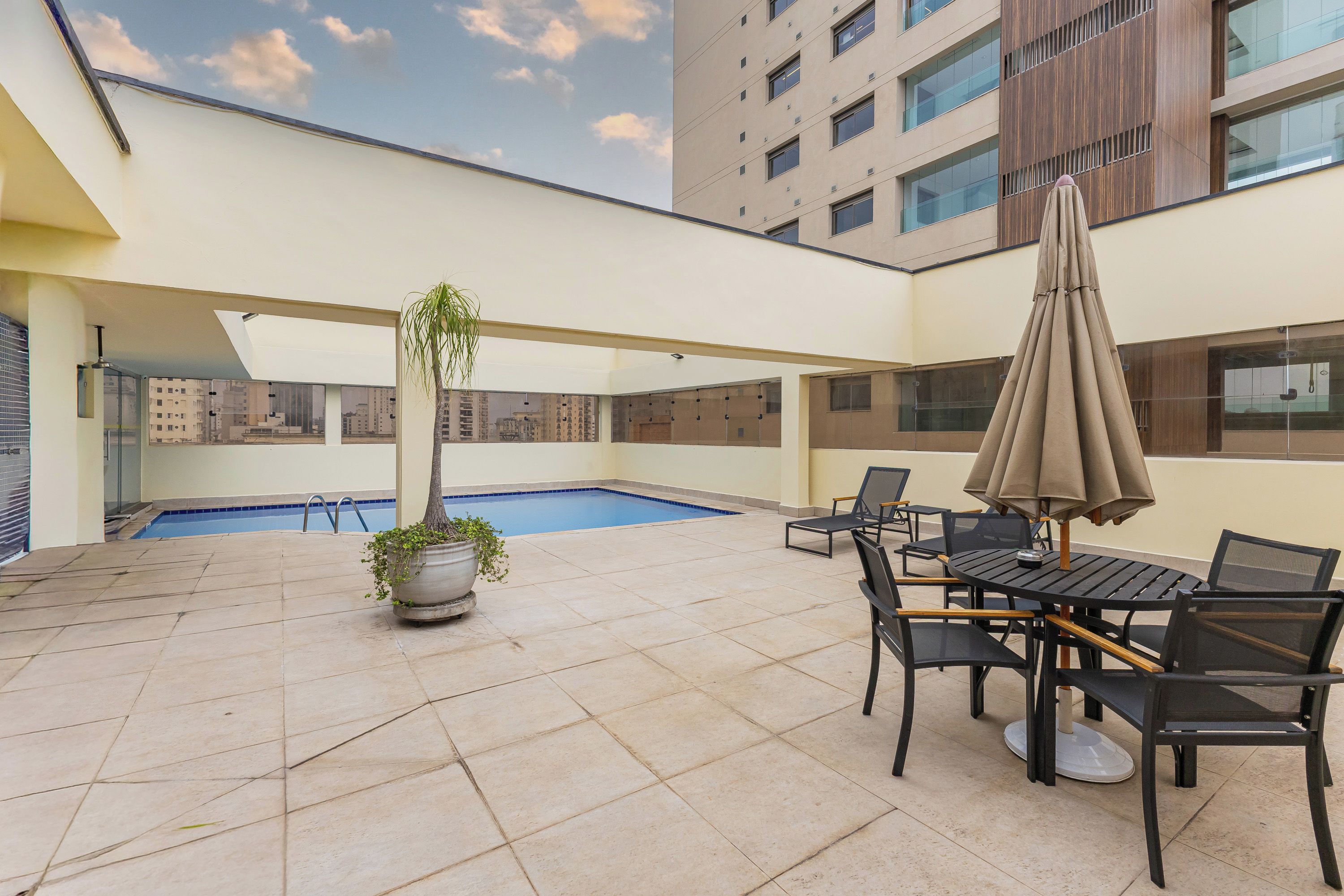 Área de piscina no terraço com mesa, cadeiras, planta em vaso e um grande guarda-sol.