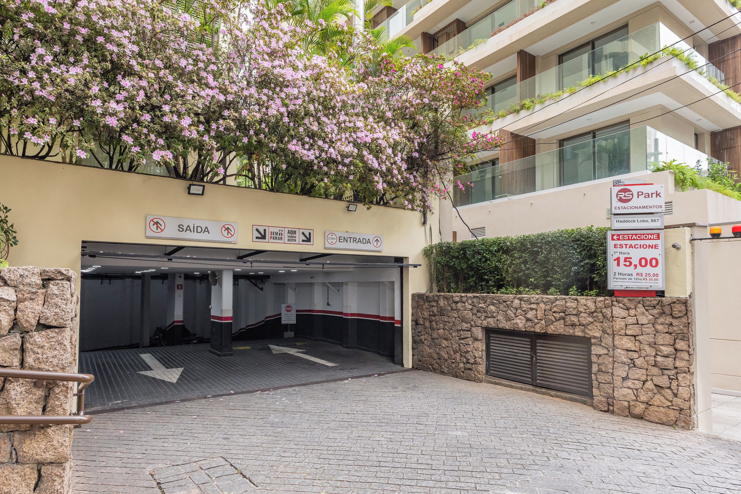 Entrada de um estacionamento com placas de sinalização, um muro de pedra e um edifício com varandas.