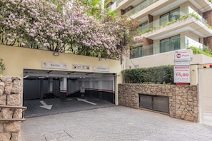 Entrada a un estacionamiento con señales de dirección, un muro de piedra y un edificio con balcones.