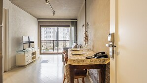 Un comedor con una mesa preparada para dos personas, vista a rascacielos a través de amplias ventanas, y una cocina moderna con un refrigerador y un aparador de vidrio.