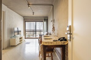 A dining area with a table set for two, a view of high-rise buildings through large windows, and a modern kitchen area with a refrigerator and a glass cabinet. - Matiz Manhattan Hotel - Gym Pool & Sauna (São Paulo)