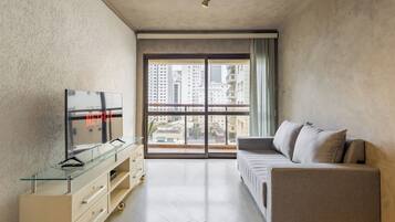 Una sala moderna con un sofá, un televisor en un mueble de vidrio y un ventanal con vista a la ciudad.