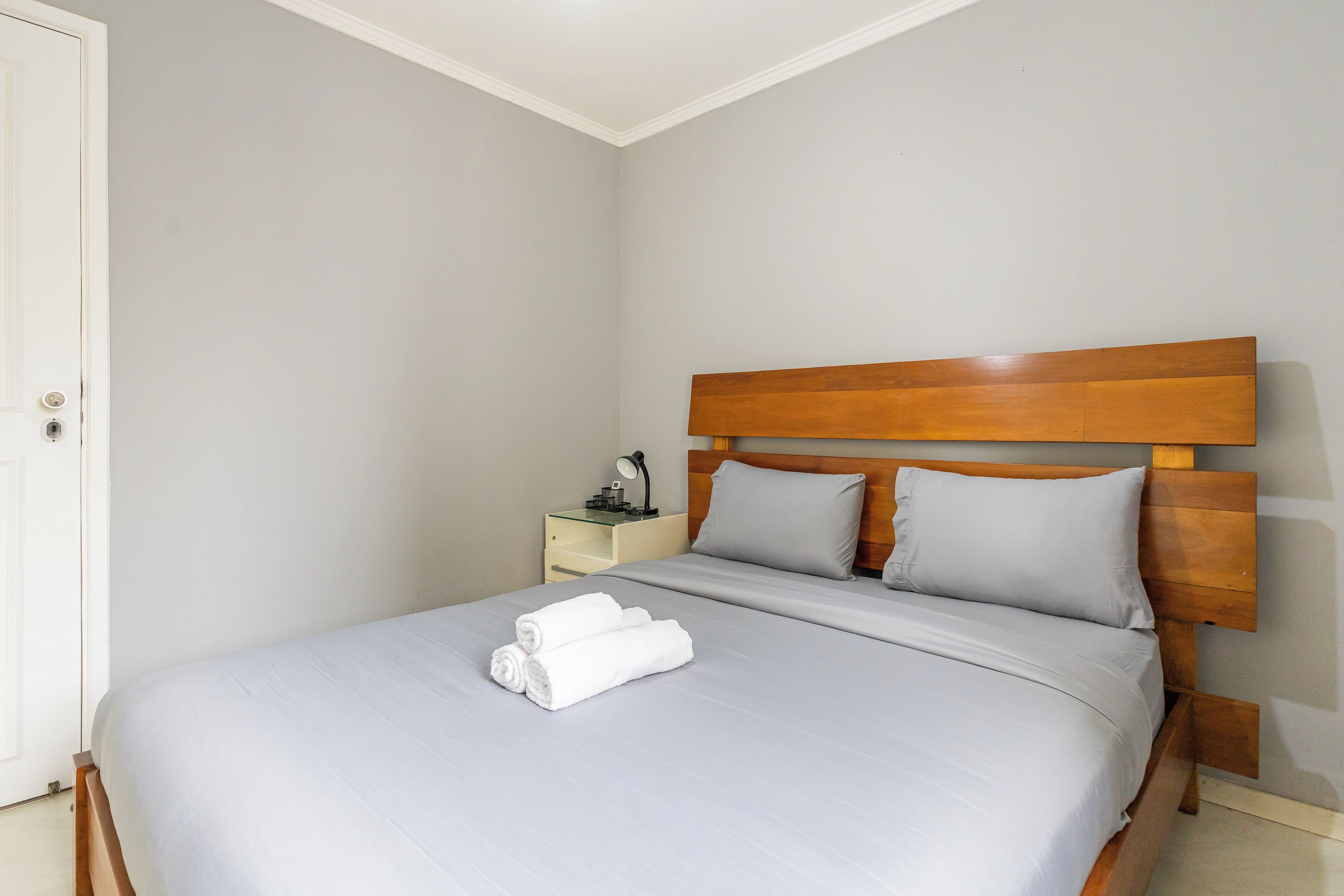 A single-bed room with a wooden headboard, a bedside table with a lamp, and a neatly made bed with rolled white towels.