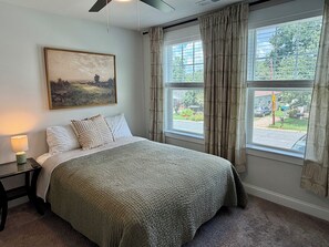 A bedroom with a bed, a nightstand, a lamp, a painting, and a window with curtains. - Fully Furnished Retreat In The Heart Of Charlotte (Charlotte)