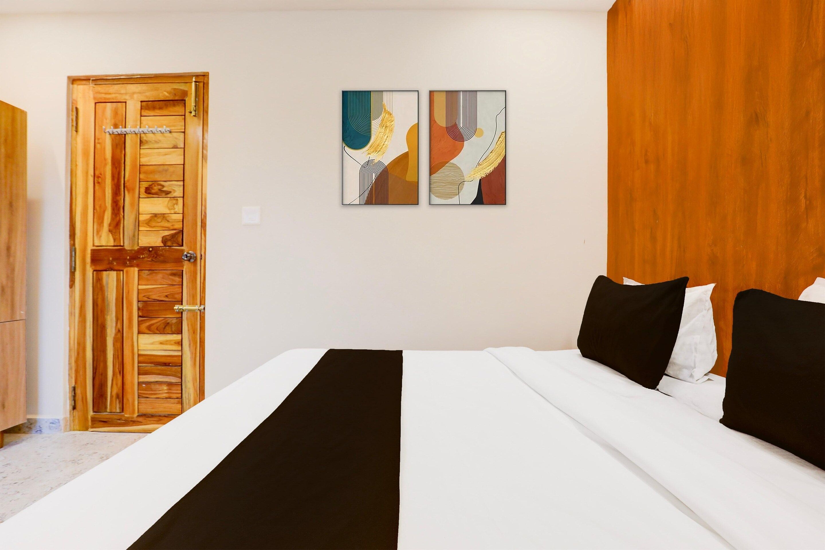 A hotel room with a wooden door, a bed with white and black bedding, and two abstract paintings on the wall.