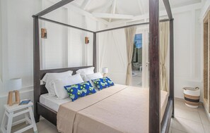 Uma cama com dossel coberta por lençóis brancos e almofadas azuis estampadas, uma mesa de cabeceira com abajur, e vista para o exterior por uma porta de vidro.