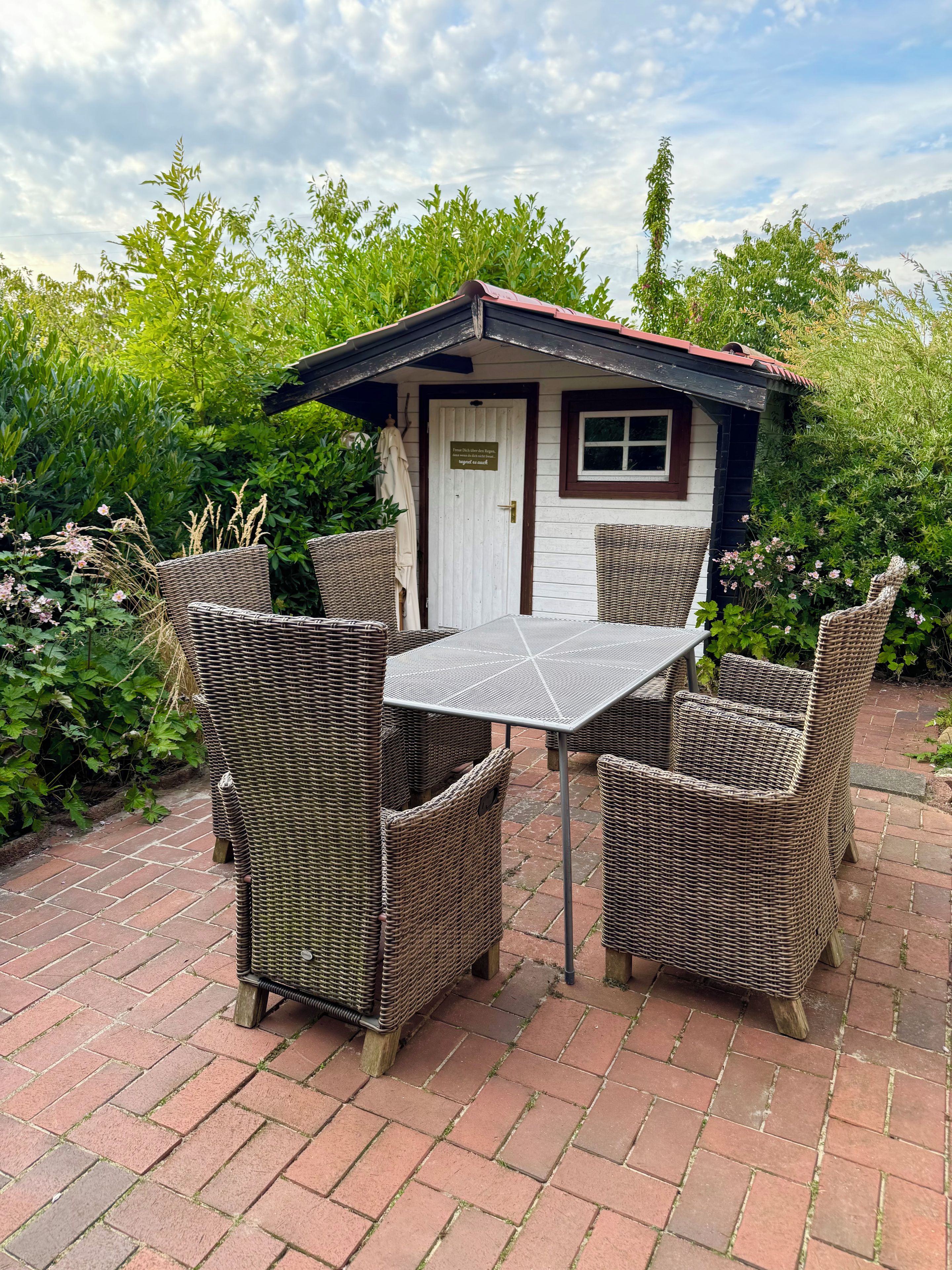 A small outdoor seating area with wicker furniture and a table, surrounded by lush greenery.