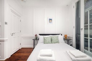 A hotel room with a large bed, a nightstand, a framed picture on the wall, and a window with blinds. - Prime London Studio | Minutes from Oxford Street (London)