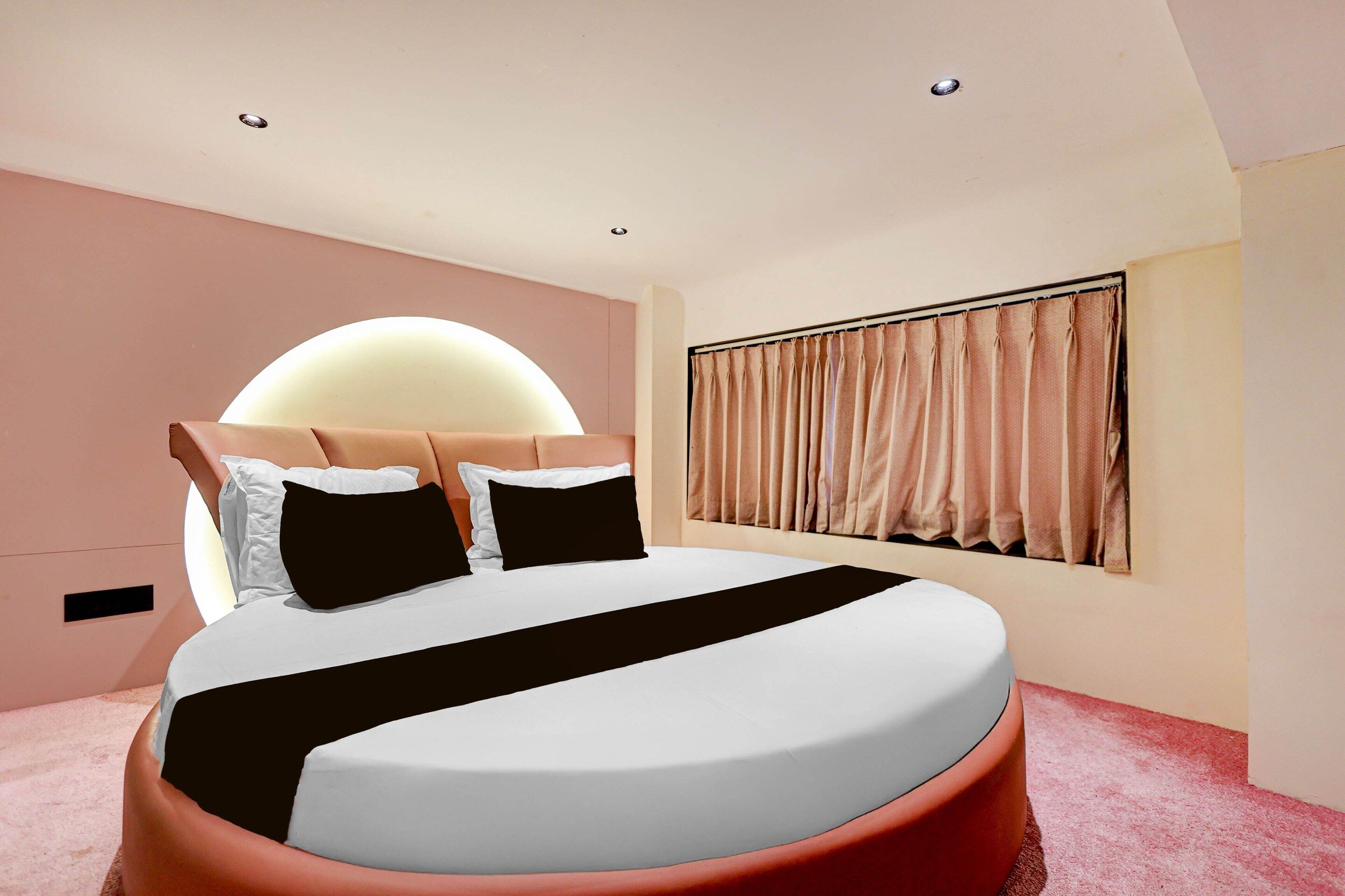 Un dormitorio moderno con una cama redonda, cabecera, ventana con cortinas y alfombra rosa.