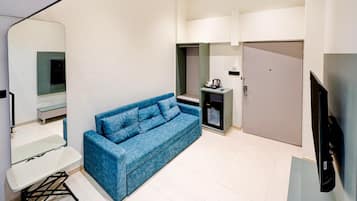 A compact living space with a blue sofa, a small table, a kitchenette with a coffee machine, and a TV.