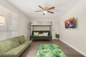 A room with a bunk bed, a green sofa, a ceiling fan, a TV mounted on the wall, and a potted plant.