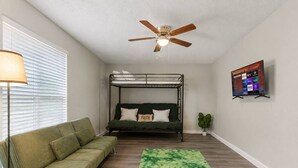 A room with a bunk bed, a sofa, a green rug, a ceiling fan, and a TV on the wall.