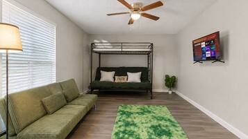 A room with a bunk bed, a sofa, a green rug, a ceiling fan, and a TV on the wall.
