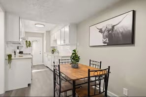 A modern kitchen with white cabinetry, a wooden dining table, and a black and white artwork on the wall.