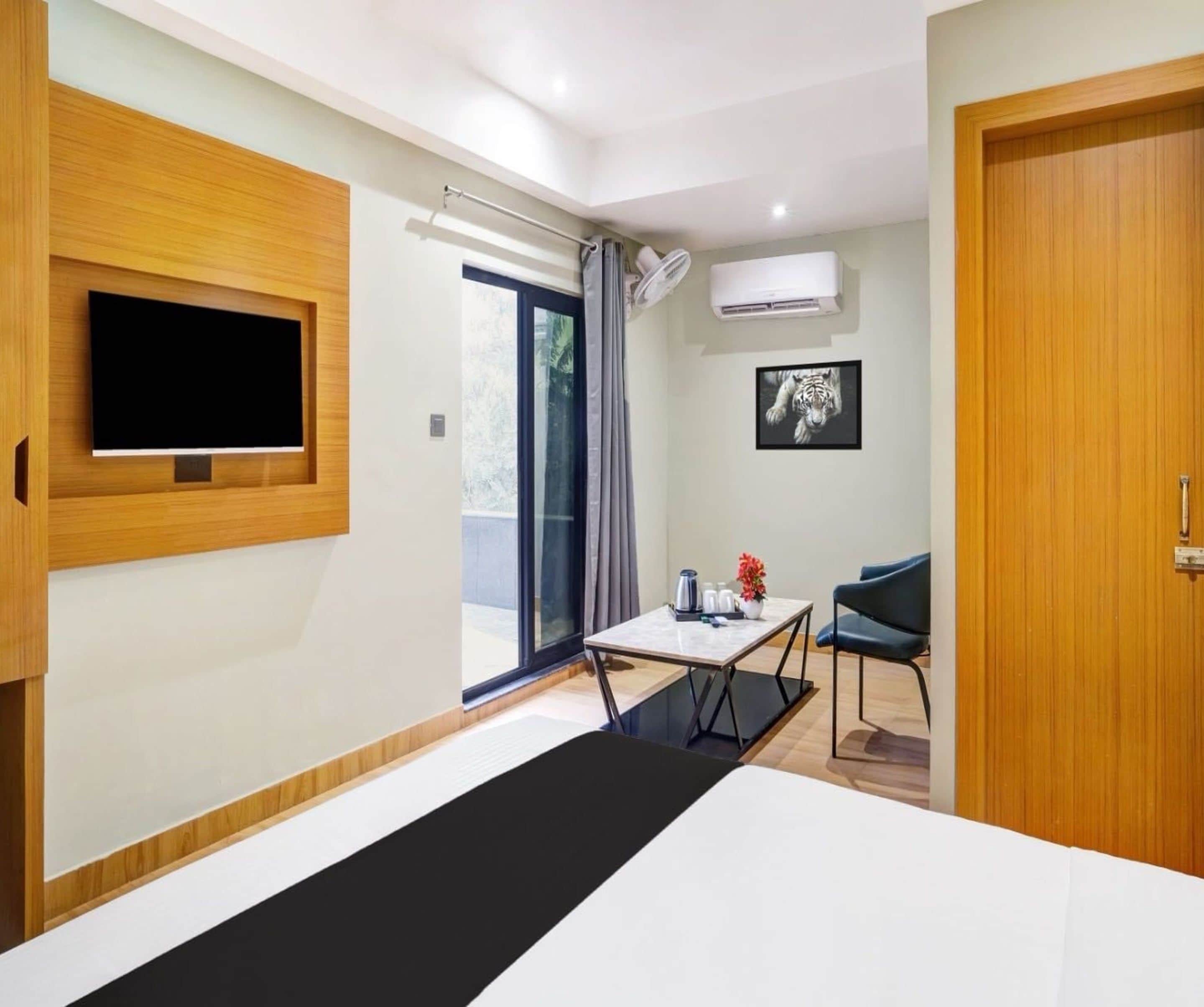 A modern hotel room with a bed, a flat-screen TV mounted on the wall, a small table with a chair, and a door.