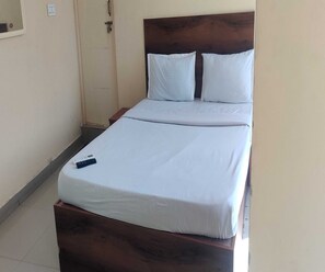 A single bed with a wooden headboard and white bedding in a room with a tiled floor and a door. - Deluxe Room RTO Office Krishnarajapura (Bengaluru)