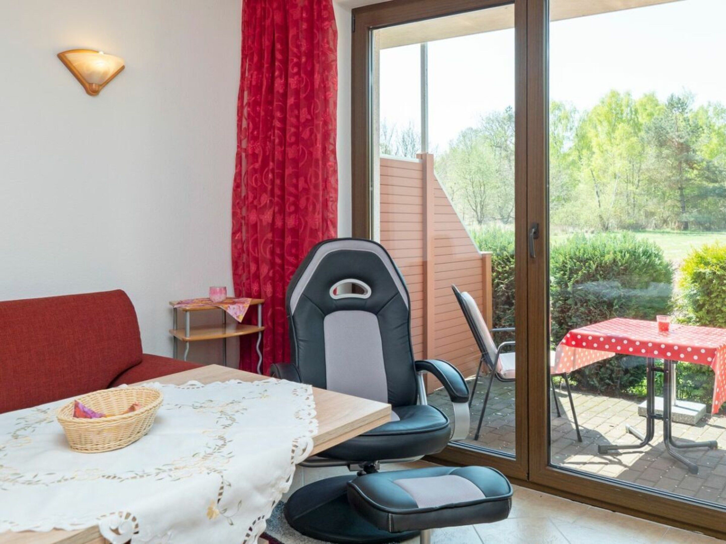 A room with a sofa, a table with a tablecloth, a chair, a window with a view of greenery, and a red curtain.