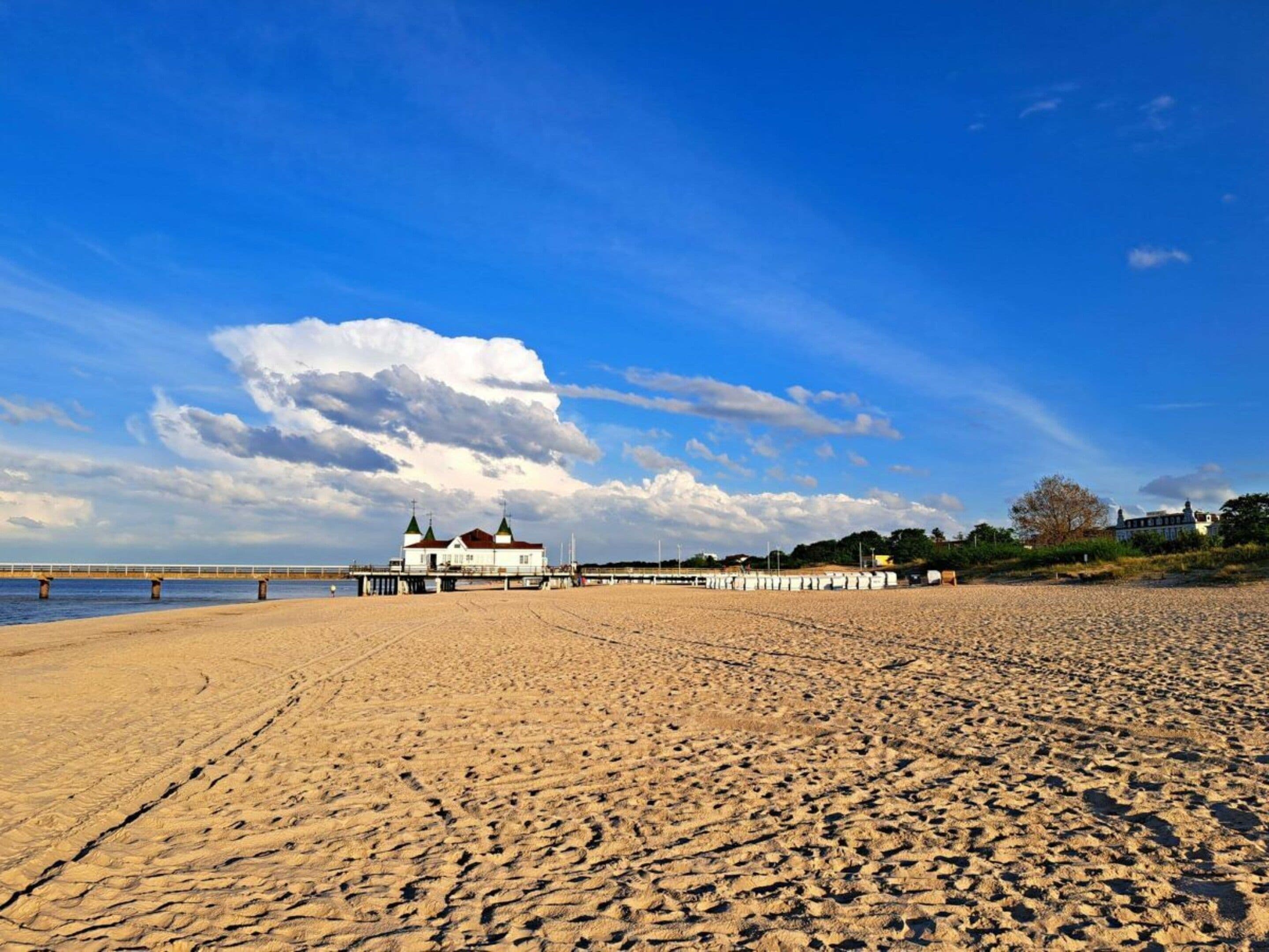 Une plage avec un bâtiment blanc surmonté de toits verts, une jetée qui s’avance dans l’eau et un ciel dégagé parsemé de nuages.
