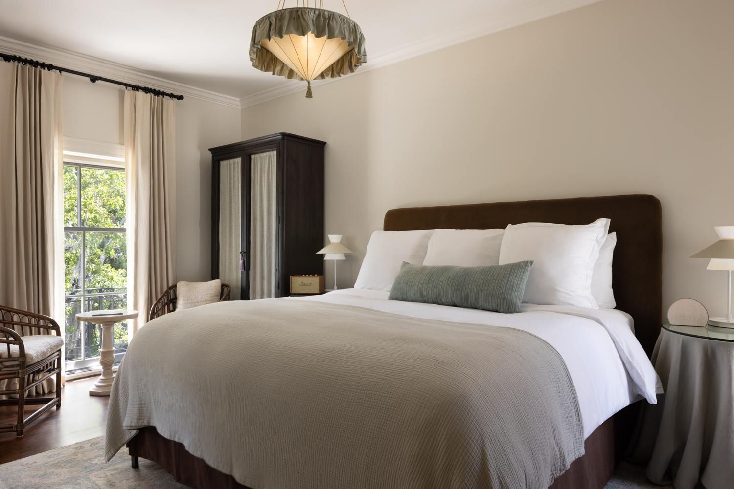 A hotel room with a large bed, a nightstand, a chair, a wardrobe, and a window with curtains.