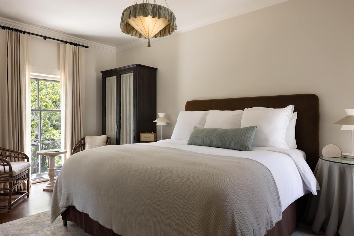 Une chambre d’hôtel avec un grand lit, une table de chevet, une chaise, une armoire et une fenêtre avec des rideaux.
